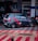 A compact hatchback car with sporty red stripes and the number '07' on its side is parked in a public area with a patterned red and gray tiled floor. The background includes a financial institution, some signage, and partially visible people.