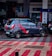 A compact hatchback car with sporty red stripes and the number '07' on its side is parked in a public area with a patterned red and gray tiled floor. The background includes a financial institution, some signage, and partially visible people.