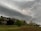 A dramatic scene of a lightning bolt striking under a dark, swirling sky. Below the stormy clouds is a landscape with a variety of trees in shades of green and brown, and an open field. In the background, there is a small wooden structure surrounded by more trees.