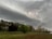 A dramatic scene of a lightning bolt striking under a dark, swirling sky. Below the stormy clouds is a landscape with a variety of trees in shades of green and brown, and an open field. In the background, there is a small wooden structure surrounded by more trees.