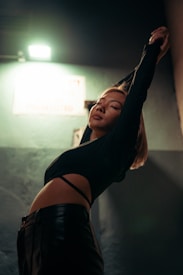 A person is captured in a dimly lit setting, stretching with eyes closed. The pose creates dynamic lines against the muted background, which includes an overhead light and an out-of-focus wall or sign. The attire includes a black top with cut-out details and black pants, contributing to a stylish and contemporary look.