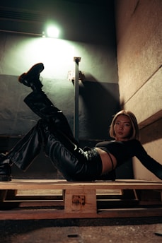 A person is lying on a wooden pallet. The setting appears to be industrial or urban, with a concrete wall and overhead lighting that creates dramatic shadows. The individual is wearing black leather clothing and boots.