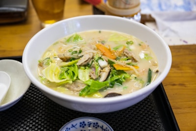 A steaming bowl of noodle soup garnished with fresh vegetables such as cabbage, carrots, and green onions, along with slices of meat and mushrooms in a creamy broth. The dish is set in a white bowl on a dark tray, accompanied by a small dish and spoon. A subtle, warm ambiance is created by a wooden table and soft lighting.
