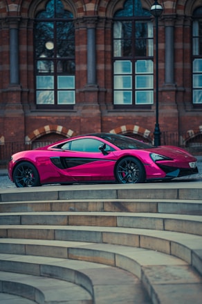 A sleek pink sports car parked in front of a historic brick building with large windows. The car is positioned on a curved stone platform, showcasing its modern and stylish design against the classic architecture.