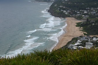 A coastal landscape includes a vast sandy beach along the ocean with waves crashing against the shore, visible from a high vantage point. The right side of the image shows a residential area with houses and greenery spread across the hills. Several people are visible on the beach, enjoying the scenery.