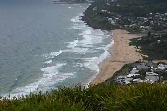 A coastal landscape includes a vast sandy beach along the ocean with waves crashing against the shore, visible from a high vantage point. The right side of the image shows a residential area with houses and greenery spread across the hills. Several people are visible on the beach, enjoying the scenery.