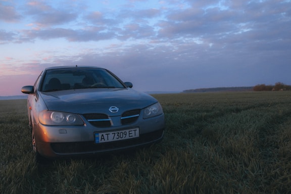 A silver car with Ukrainian license plates is parked in the middle of a green field under a partly cloudy sky during sunrise or sunset. The sky has a warm, colorful gradient effect with shades of blue, pink, and orange.