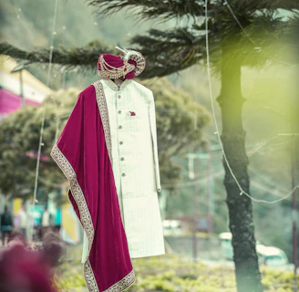 A traditional Indian men's outfit is displayed on a mannequin in an outdoor setting. The attire consists of a cream-colored sherwani with intricate designs, accompanied by a maroon and gold embroidered turban and scarf. The background features trees and a blurred natural landscape.