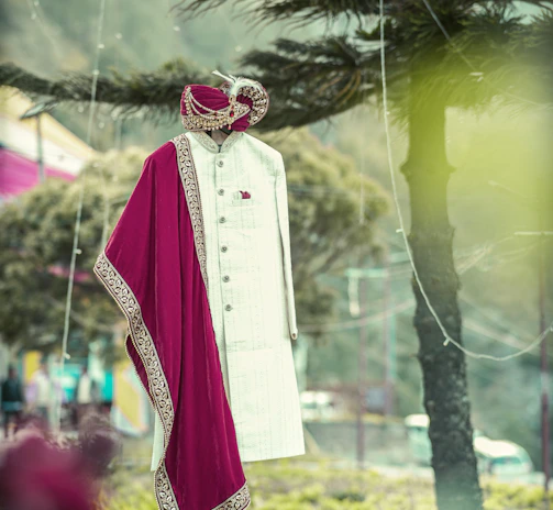 A traditional Indian men's outfit is displayed on a mannequin in an outdoor setting. The attire consists of a cream-colored sherwani with intricate designs, accompanied by a maroon and gold embroidered turban and scarf. The background features trees and a blurred natural landscape.