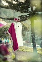 A traditional Indian men's outfit is displayed on a mannequin in an outdoor setting. The attire consists of a cream-colored sherwani with intricate designs, accompanied by a maroon and gold embroidered turban and scarf. The background features trees and a blurred natural landscape.