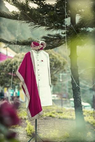 A traditional Indian men's outfit is displayed on a mannequin in an outdoor setting. The attire consists of a cream-colored sherwani with intricate designs, accompanied by a maroon and gold embroidered turban and scarf. The background features trees and a blurred natural landscape.