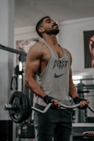 A muscular individual wearing a sleeveless shirt performs a barbell curl exercise in a gym. The shirt has the word 'BEAST' and a logo on it. The person is using a barbell with weighted plates, and wears wrist straps for support. Gym equipment and posters are visible in the background.