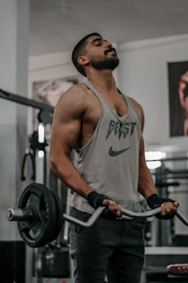 A muscular individual wearing a sleeveless shirt performs a barbell curl exercise in a gym. The shirt has the word 'BEAST' and a logo on it. The person is using a barbell with weighted plates, and wears wrist straps for support. Gym equipment and posters are visible in the background.