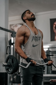 A muscular individual wearing a sleeveless shirt performs a barbell curl exercise in a gym. The shirt has the word 'BEAST' and a logo on it. The person is using a barbell with weighted plates, and wears wrist straps for support. Gym equipment and posters are visible in the background.