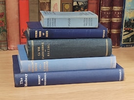 A stack of vintage hardcover books is neatly arranged on a wooden surface. The spines of the books have a variety of colors including blue, teal, and dark red. In the background, more books are visible on a shelf, adding an aesthetic of a classical library setting.