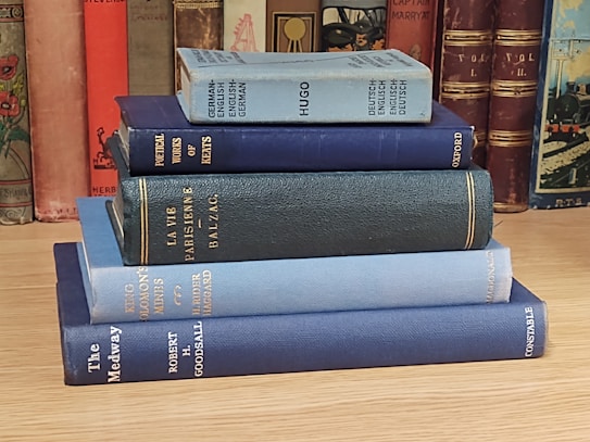 A stack of vintage hardcover books is neatly arranged on a wooden surface. The spines of the books have a variety of colors including blue, teal, and dark red. In the background, more books are visible on a shelf, adding an aesthetic of a classical library setting.