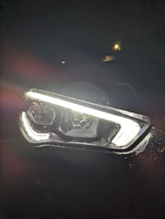 An illuminated car headlight is surrounded by darkness, creating a strong contrast. There is visible moisture or raindrops around the edges of the headlight. A small light or reflection can be seen in the background.