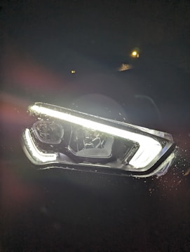 An illuminated car headlight is surrounded by darkness, creating a strong contrast. There is visible moisture or raindrops around the edges of the headlight. A small light or reflection can be seen in the background.