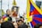 A group of people participate in a rally, with many holding flags that prominently feature red, blue, and yellow. One person in the foreground is holding a framed photograph of a person in traditional red and yellow robes, displaying a serene expression. The gathering appears to be outdoors, with attendees looking attentive and focused.