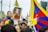 A group of people participate in a rally, with many holding flags that prominently feature red, blue, and yellow. One person in the foreground is holding a framed photograph of a person in traditional red and yellow robes, displaying a serene expression. The gathering appears to be outdoors, with attendees looking attentive and focused.