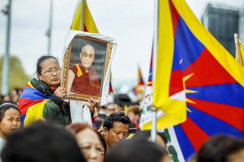 A group of people participate in a rally, with many holding flags that prominently feature red, blue, and yellow. One person in the foreground is holding a framed photograph of a person in traditional red and yellow robes, displaying a serene expression. The gathering appears to be outdoors, with attendees looking attentive and focused.