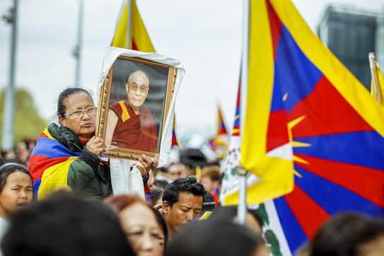 A group of people participate in a rally, with many holding flags that prominently feature red, blue, and yellow. One person in the foreground is holding a framed photograph of a person in traditional red and yellow robes, displaying a serene expression. The gathering appears to be outdoors, with attendees looking attentive and focused.