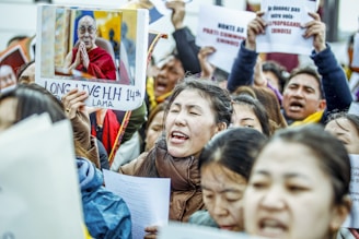 A group of protestors, possibly Tibetan, are gathered together holding signs and pictures. One prominent sign features an image of a man in traditional robes with a message of support. Several people are singing or chanting passionately.