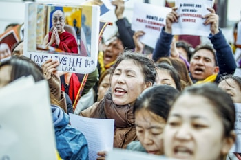 A group of protestors, possibly Tibetan, are gathered together holding signs and pictures. One prominent sign features an image of a man in traditional robes with a message of support. Several people are singing or chanting passionately.