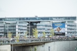 A modern glass office building with the logo 'france.tv' prominently displayed at the top. A billboard featuring a person and the text 'BARDOT' is visible. In front of the building, there is a large crowd of people on a bridge or walkway, along with several small trees.