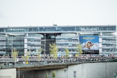 A modern glass office building with the logo 'france.tv' prominently displayed at the top. A billboard featuring a person and the text 'BARDOT' is visible. In front of the building, there is a large crowd of people on a bridge or walkway, along with several small trees.