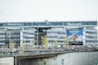 A modern glass office building with the logo 'france.tv' prominently displayed at the top. A billboard featuring a person and the text 'BARDOT' is visible. In front of the building, there is a large crowd of people on a bridge or walkway, along with several small trees.