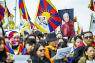 A large group of people gathered, many holding Tibetan flags. The crowd includes individuals wearing traditional Tibetan attire and facial coverings with similar flag designs. A prominent poster of a monk in red robes is visible among the flags.