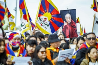 A large group of people gathered, many holding Tibetan flags. The crowd includes individuals wearing traditional Tibetan attire and facial coverings with similar flag designs. A prominent poster of a monk in red robes is visible among the flags.