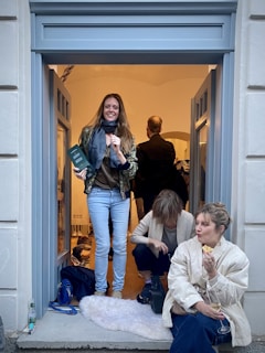 Three people are interacting in an indoor space framed by a light blue doorway. One person stands holding a drink, smiling, while the other two are sitting on the ground, each engaged in personal activities. One of the seated individuals is eating and holding a glass, while the other appears to adjust their shoe. The room is warmly lit, and the wall colors are neutral, contrasting with the cooler tones of the door frame.