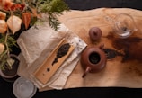 A wooden surface is adorned with a bamboo tray holding loose tea leaves, accompanied by a brown clay teapot with its lid beside it. A transparent glass pitcher and a decorative paper with Chinese characters are also present. Sprigs of green foliage and orange lantern-like flower decorations encroach from the upper left corner, adding an organic touch.