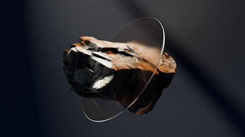 A large, rough, dark-colored rock is suspended in mid-air, intersected by a thin, transparent circular disc. The rock has various textures and shades of brown and gray, highlighted by focused lighting that accentuates its irregular surface.