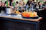 A bar setting with a wooden countertop featuring a variety of liquor bottles and a mixed drink station. In the foreground, a tray holds four vibrant cocktails, garnished with orange slices. The background is bustling with people.