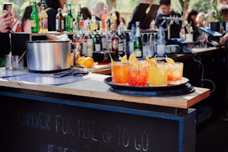 A bar setting with a wooden countertop featuring a variety of liquor bottles and a mixed drink station. In the foreground, a tray holds four vibrant cocktails, garnished with orange slices. The background is bustling with people.