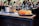 A bar setting with a wooden countertop featuring a variety of liquor bottles and a mixed drink station. In the foreground, a tray holds four vibrant cocktails, garnished with orange slices. The background is bustling with people.