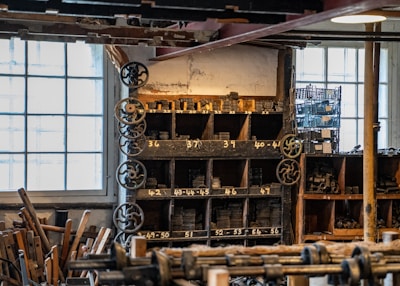 An interior of a workshop filled with wooden shelves holding numerous metal gears and parts. Several large windows allow natural light to illuminate the space, highlighting both the aged and industrial aesthetic. Gears and machinery components are neatly organized in compartments labeled with numbers. Wooden tools and equipment are scattered throughout, contributing to a cluttered yet orderly atmosphere.