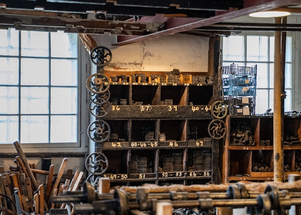 An interior of a workshop filled with wooden shelves holding numerous metal gears and parts. Several large windows allow natural light to illuminate the space, highlighting both the aged and industrial aesthetic. Gears and machinery components are neatly organized in compartments labeled with numbers. Wooden tools and equipment are scattered throughout, contributing to a cluttered yet orderly atmosphere.