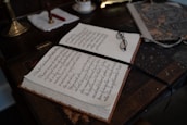 An open book with neatly handwritten text lies on a wooden desk. A pair of eyeglasses rests on the book, beside a ribbon bookmark. The desk also holds a marbled folder and a brass lamp with a white cloth and a red wax seal next to it.