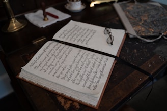 An open book with neatly handwritten text lies on a wooden desk. A pair of eyeglasses rests on the book, beside a ribbon bookmark. The desk also holds a marbled folder and a brass lamp with a white cloth and a red wax seal next to it.