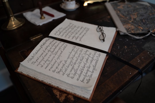 An open book with neatly handwritten text lies on a wooden desk. A pair of eyeglasses rests on the book, beside a ribbon bookmark. The desk also holds a marbled folder and a brass lamp with a white cloth and a red wax seal next to it.