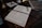 An open book with neatly handwritten text lies on a wooden desk. A pair of eyeglasses rests on the book, beside a ribbon bookmark. The desk also holds a marbled folder and a brass lamp with a white cloth and a red wax seal next to it.