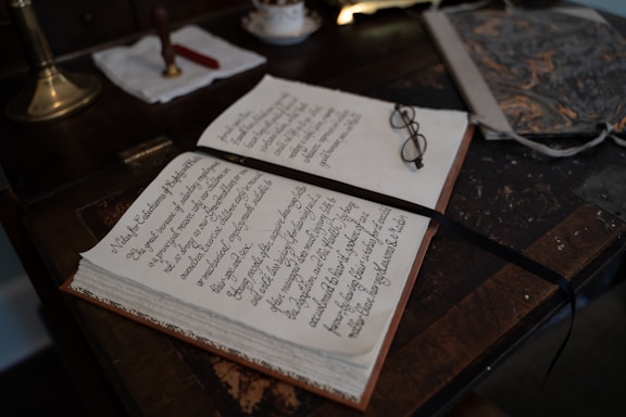 An open book with neatly handwritten text lies on a wooden desk. A pair of eyeglasses rests on the book, beside a ribbon bookmark. The desk also holds a marbled folder and a brass lamp with a white cloth and a red wax seal next to it.
