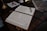 An open book with neatly handwritten text lies on a wooden desk. A pair of eyeglasses rests on the book, beside a ribbon bookmark. The desk also holds a marbled folder and a brass lamp with a white cloth and a red wax seal next to it.