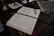 An open book with neatly handwritten text lies on a wooden desk. A pair of eyeglasses rests on the book, beside a ribbon bookmark. The desk also holds a marbled folder and a brass lamp with a white cloth and a red wax seal next to it.