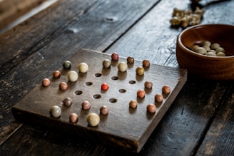 A wooden solitaire board with multiple small, colorful marbles positioned in holes. The board has a grid-like pattern, and there is an empty space in the center. A wooden bowl containing additional marbles is placed to the side on the rough-textured wooden surface.