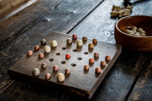 A wooden solitaire board with multiple small, colorful marbles positioned in holes. The board has a grid-like pattern, and there is an empty space in the center. A wooden bowl containing additional marbles is placed to the side on the rough-textured wooden surface.